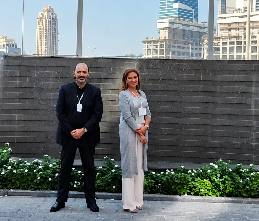 Two professionals wearing ID badges stand against a gray brick wall with green plants and a city skyline in the background