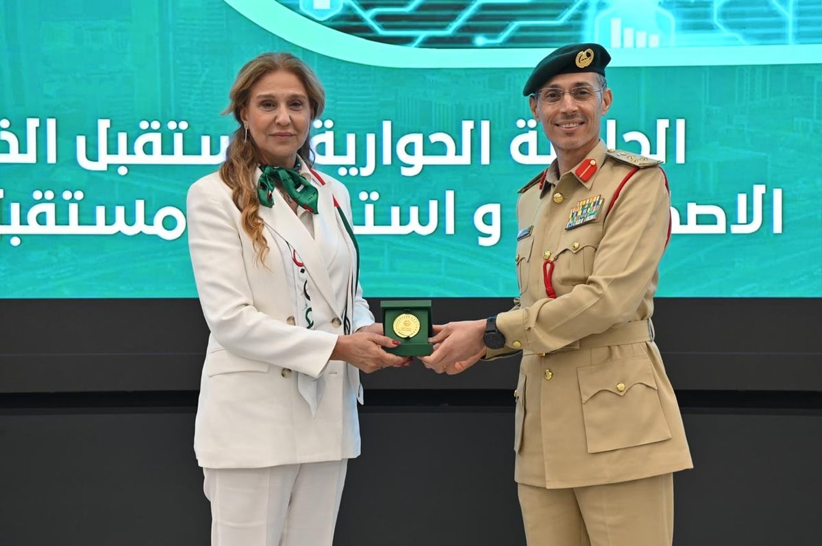 Two officials exchange a plaque during a formal ceremony with Arabic text displayed on screen behind them.